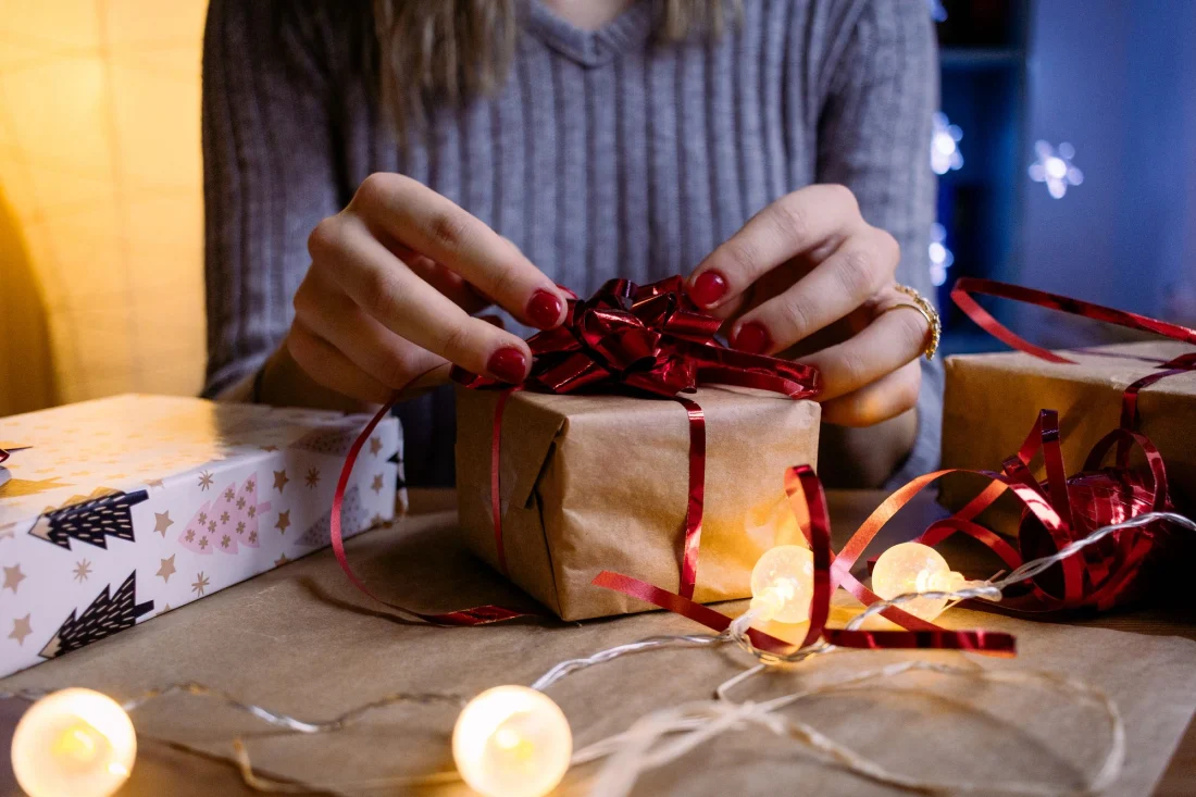 Hände binden eine rote Schleife um ein Geschenk, daneben eine Lichterkette