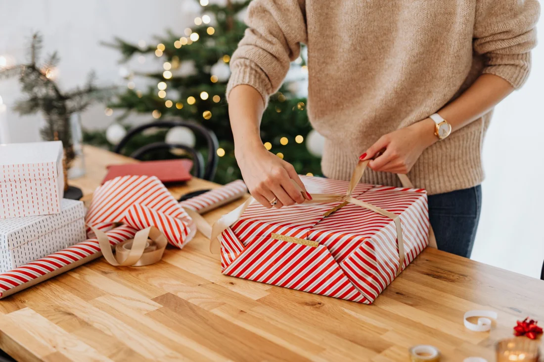 Geschenke werden auf einem Holztisch eingepackt, im Hintergrund ein Weihnachtsbaum