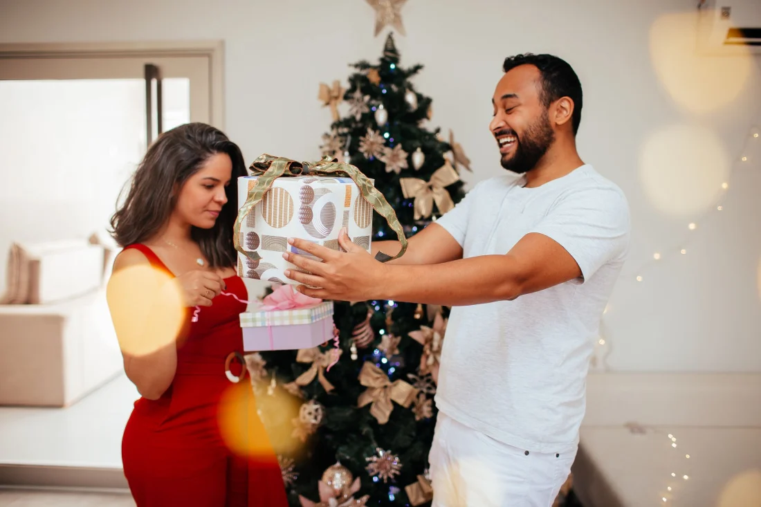 Ein Paar tauscht vor dem Weihnachtsbaum Geschenke aus, er lacht